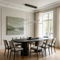 Linear pendant light made of wavy black LED bars hung from a slim rectangular canopy above an oval dining table in a panelled room.