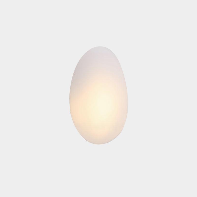 Bubble wall lamp with a rounded opal white glass shade in an egg form, lit from inside, mounted alone on a plain pale wall.