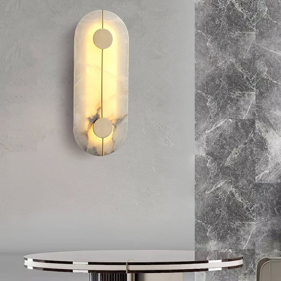 Wall light with a tall rounded alabaster panel, a slim brass rod and two brass discs lit beside a round table and a marble panel.