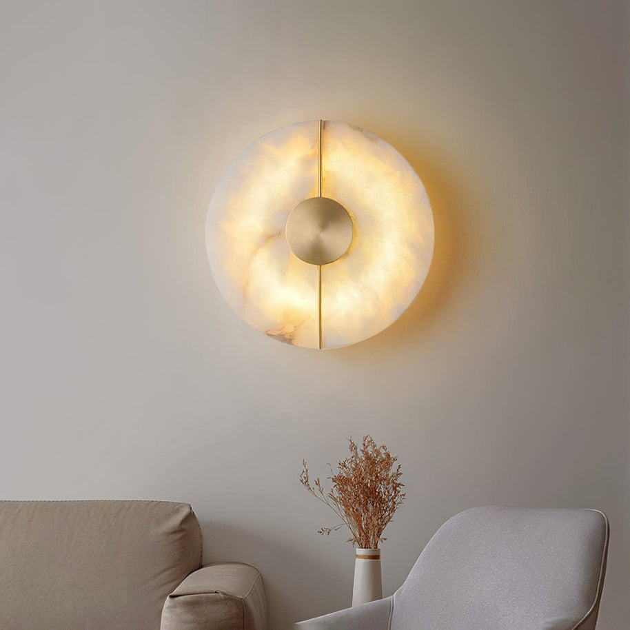 Wall light with a round veined alabaster disc, a slim brass rod and a brass centre disc lit above a sofa.