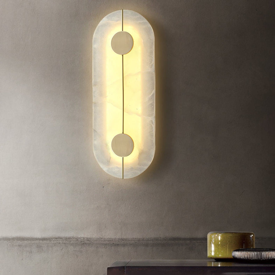 Wall light with a tall rounded alabaster panel, a slim brass rod and two brass discs lit, shown large on a plain wall.
