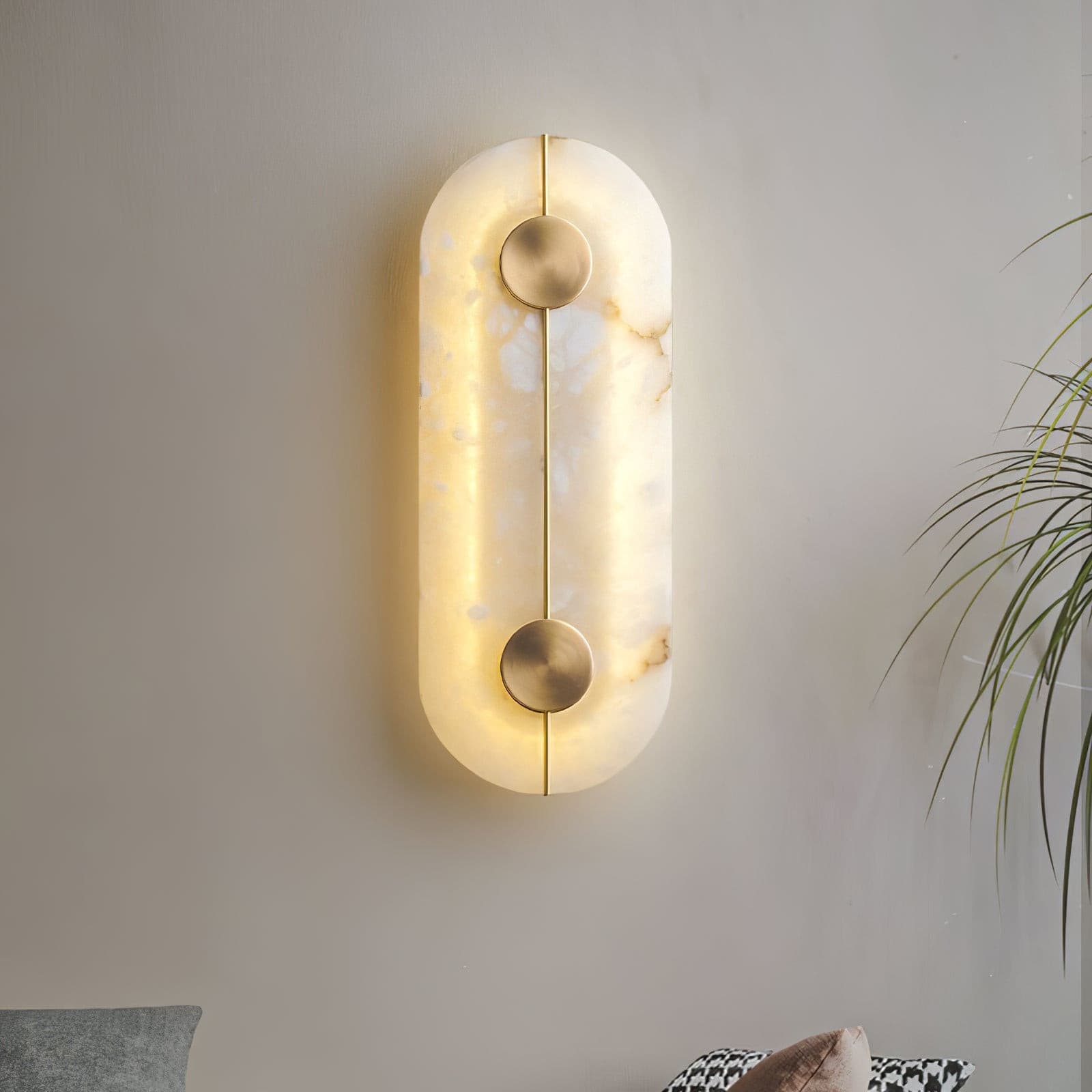 Wall light with a tall rounded alabaster panel, a slim brass rod and two brass discs lit beside a plant.