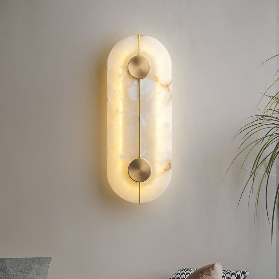 Wall light with a tall rounded alabaster panel, a slim brass rod and two brass discs lit beside a plant.