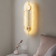 Wall light with a tall rounded alabaster panel, a slim brass rod and two brass discs lit beside a bed.