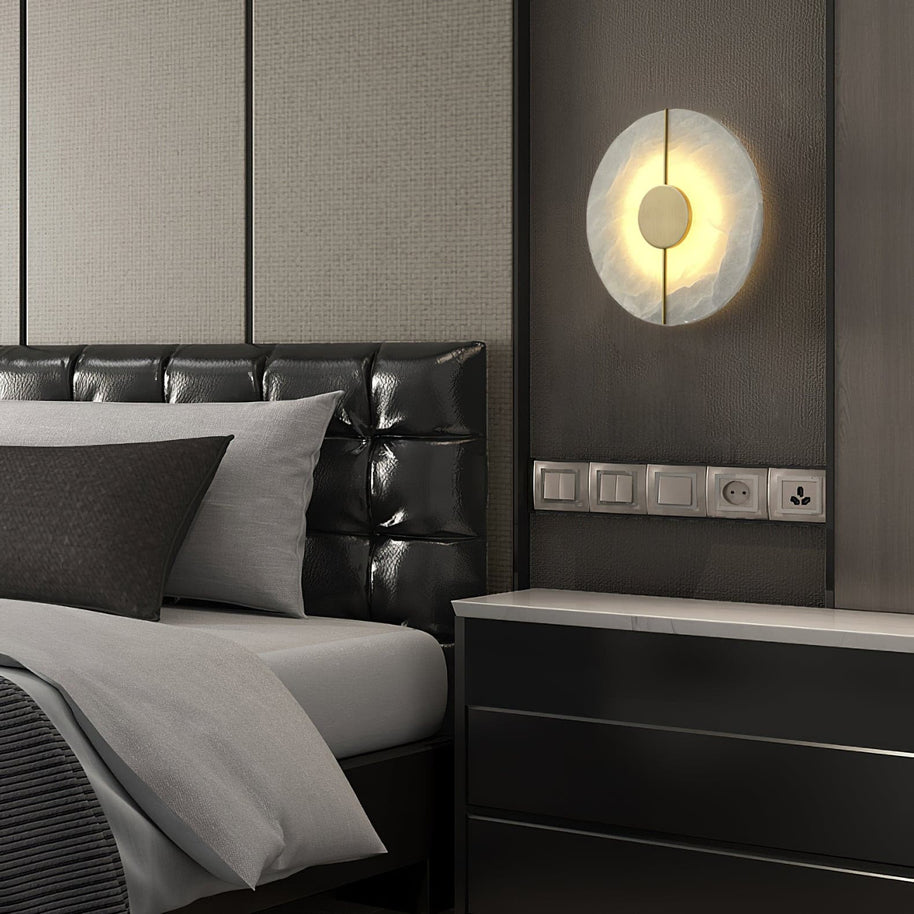 Wall light with a round veined alabaster disc, a slim brass rod and a brass centre disc lit above a bedside chest beside a leather headboard.