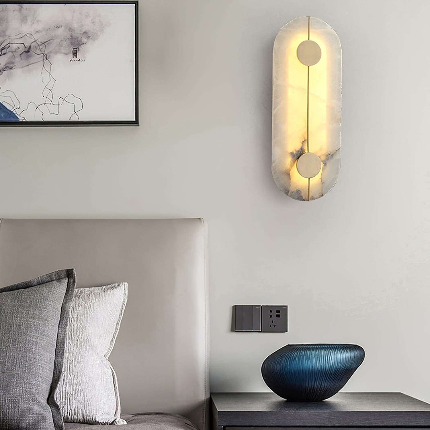 Wall light with a tall rounded alabaster panel, a slim brass rod and two brass discs lit beside a bed and a framed drawing.