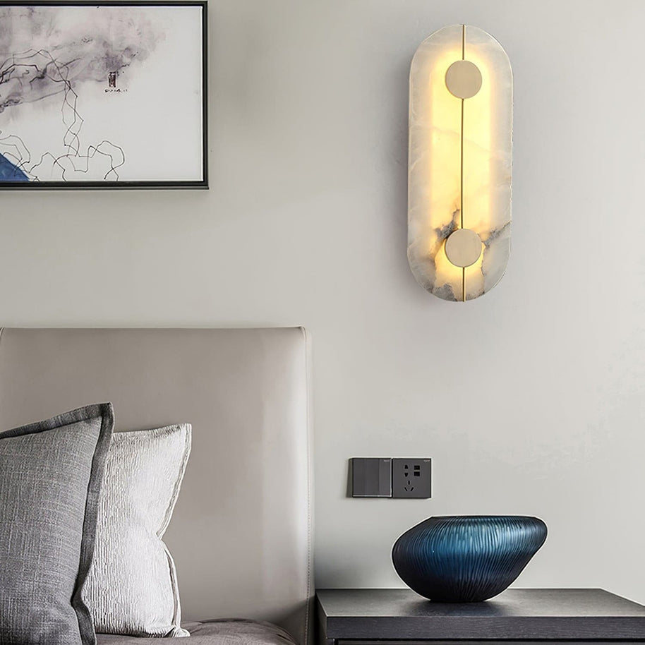 Wall light with a tall rounded alabaster panel, a slim brass rod and two brass discs lit beside a bed and a framed drawing.