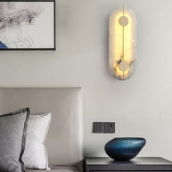 Wall light with a tall rounded alabaster panel, a slim brass rod and two brass discs lit beside a bed and a framed drawing.