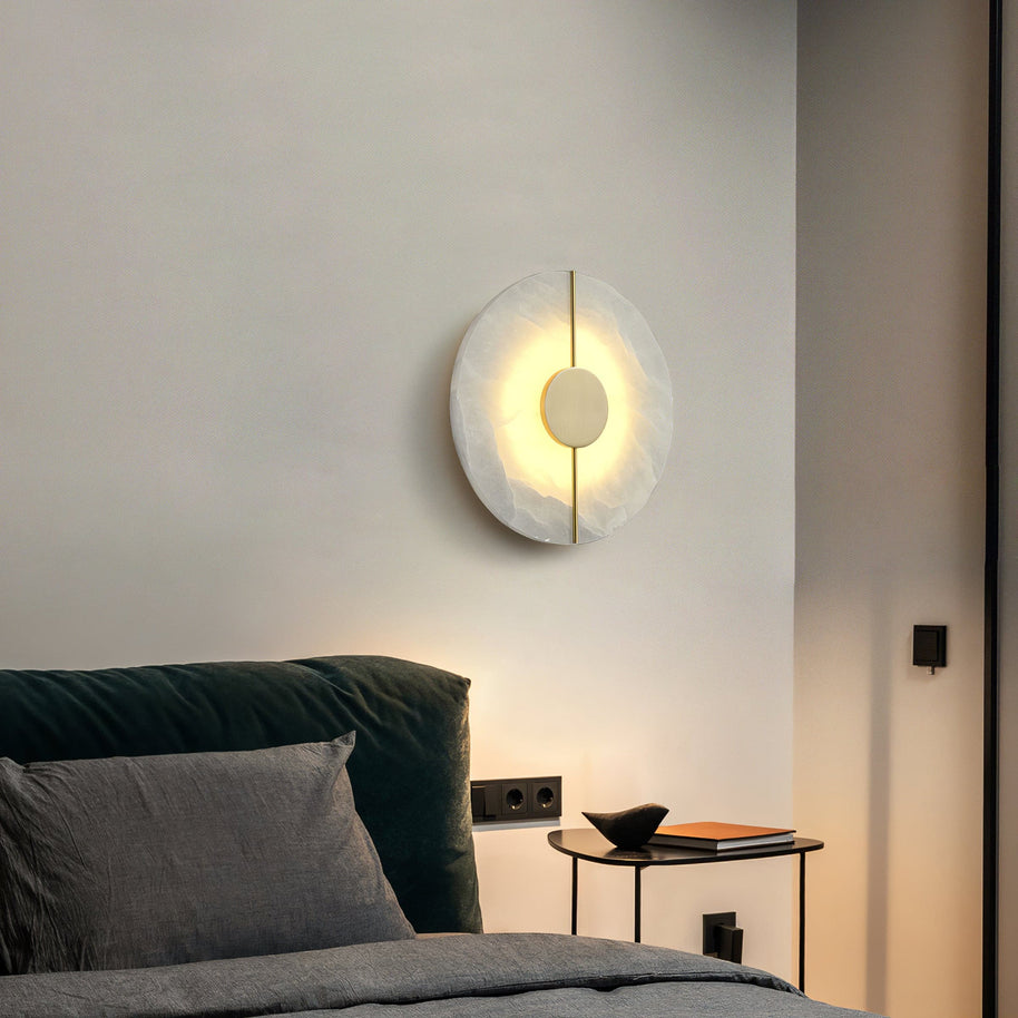 Wall light with a round veined alabaster disc, a slim brass rod and a brass centre disc lit beside a bed with a green headboard.