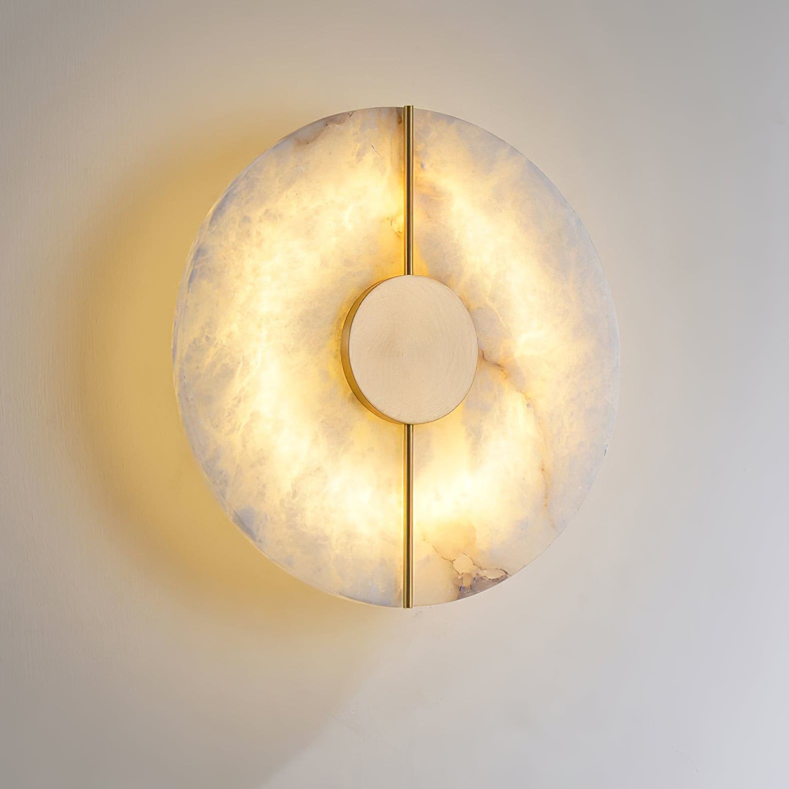 Wall light with a round veined alabaster disc, a slim brass rod and a brass centre disc, lit, shown close on a pale wall.