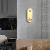 Wall light with a tall rounded alabaster panel, a slim brass rod and two brass discs lit on a plaster wall beside a kitchen.