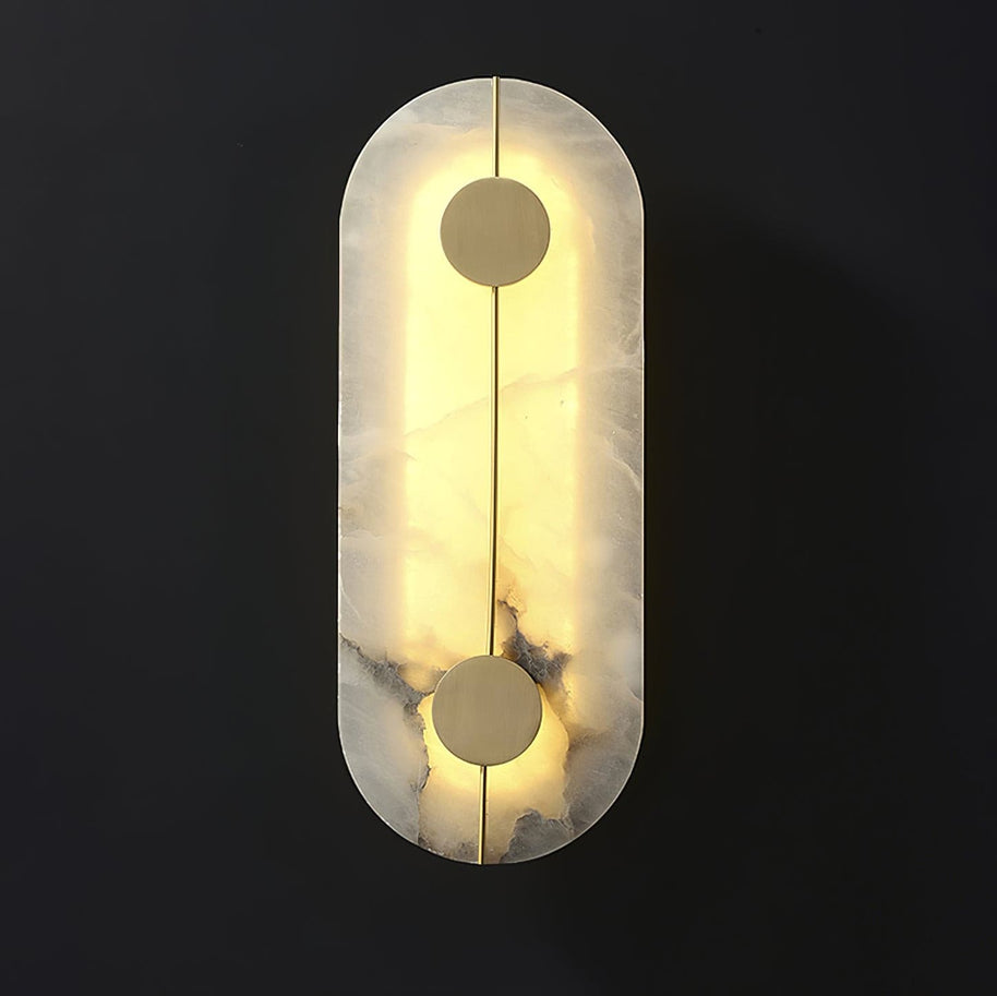 Wall light with a tall rounded alabaster panel, a slim brass rod and two brass discs lit on a dark background.