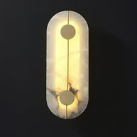 Wall light with a tall rounded alabaster panel, a slim brass rod and two brass discs lit on a dark background.