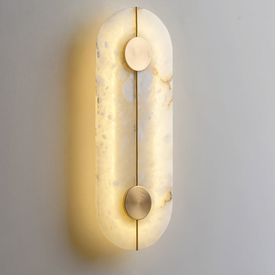 Wall light with a tall rounded alabaster panel, a slim brass rod and two brass discs, lit, shown close on a pale wall.