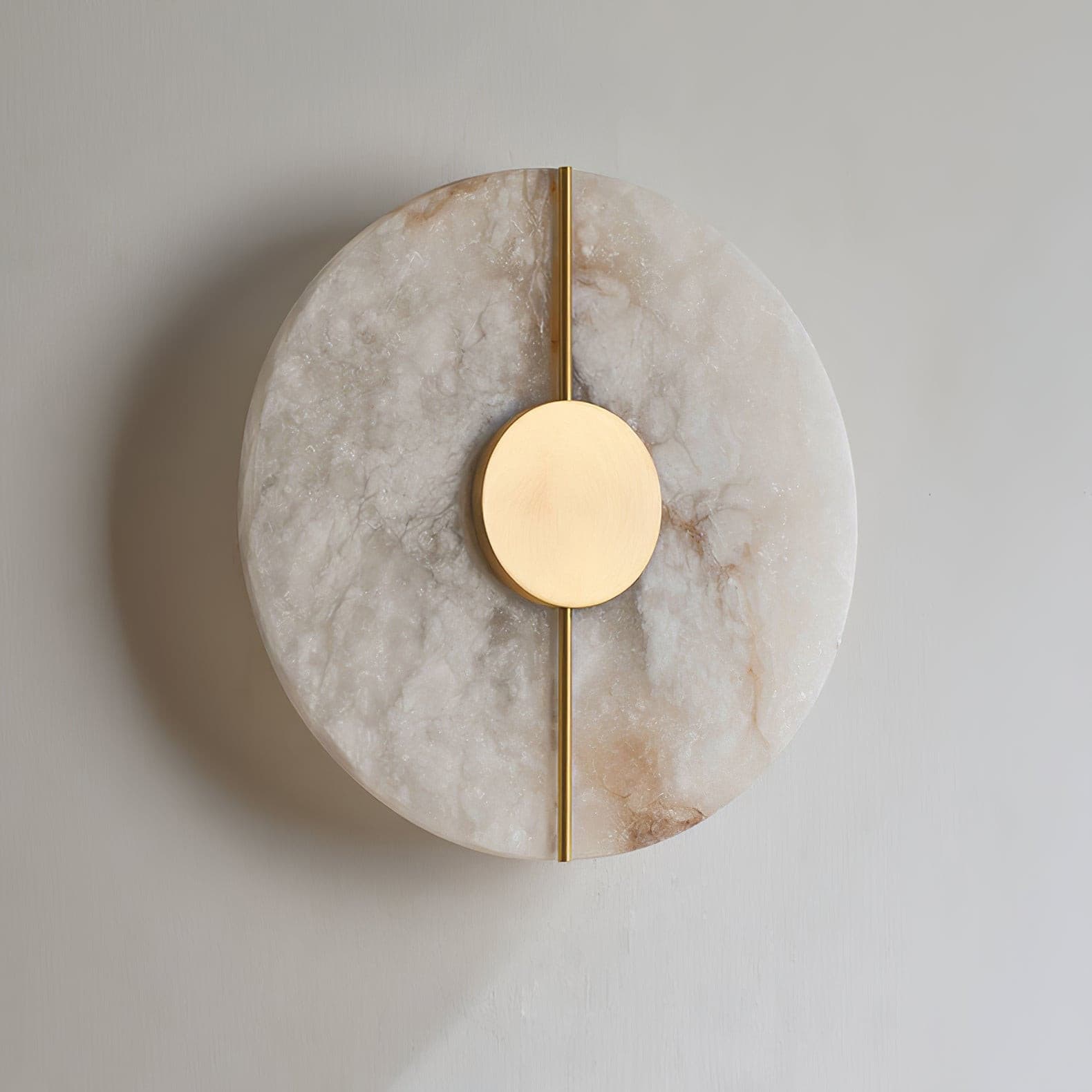 Wall light with a round veined alabaster disc, a slim brass rod and a brass centre disc, unlit, on a plaster wall.
