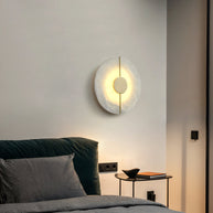 Wall light with a round veined alabaster disc, a slim brass rod and a brass centre disc lit beside a bed with a green headboard.
