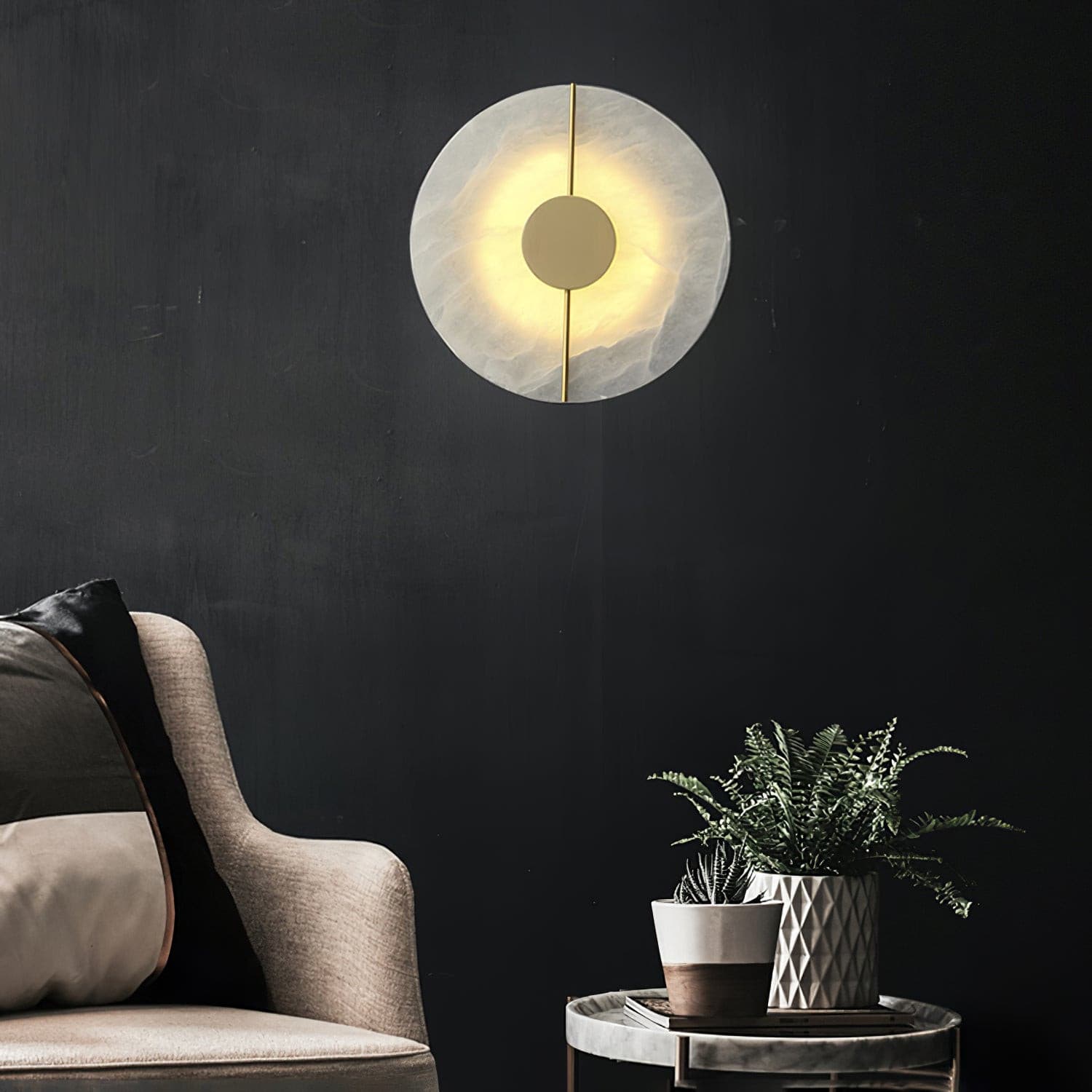 Wall light with a round veined alabaster disc, a slim brass rod and a brass centre disc lit on a dark wall beside an armchair.