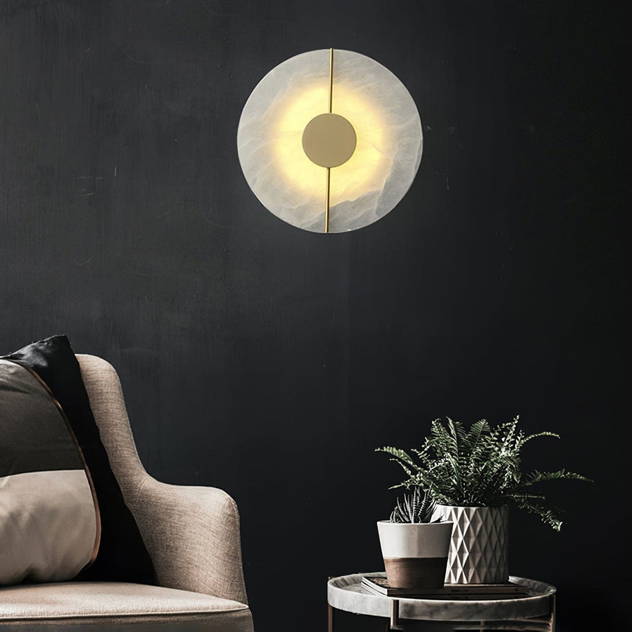 Wall light with a round veined alabaster disc, a slim brass rod and a brass centre disc lit on a dark wall beside an armchair.