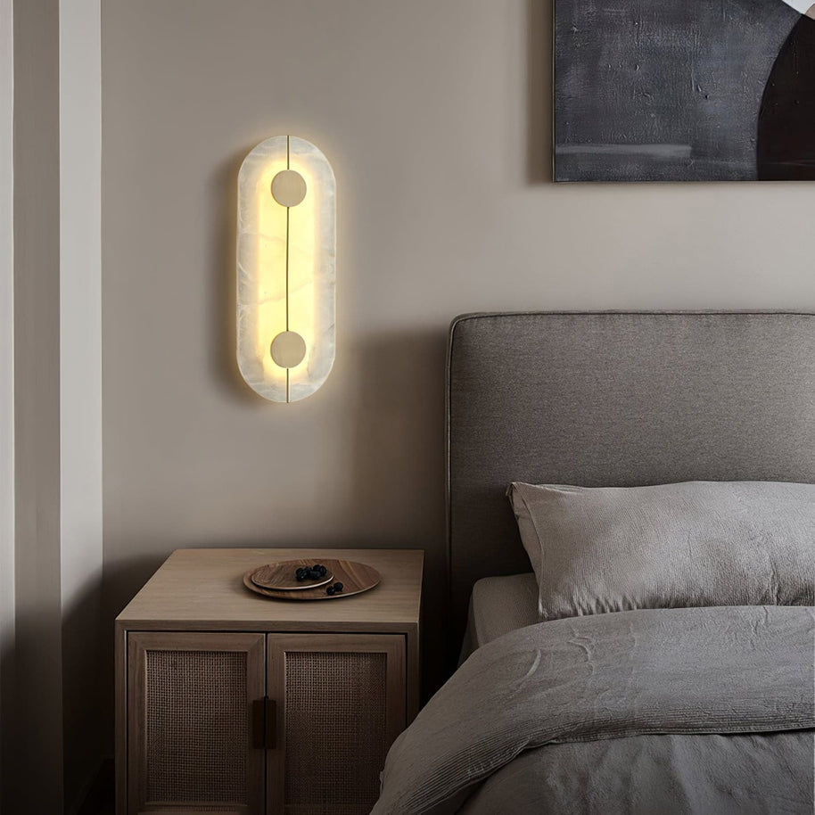 Wall light with a tall rounded alabaster panel, a slim brass rod and two brass discs lit beside a bed and a wooden bedside cabinet.