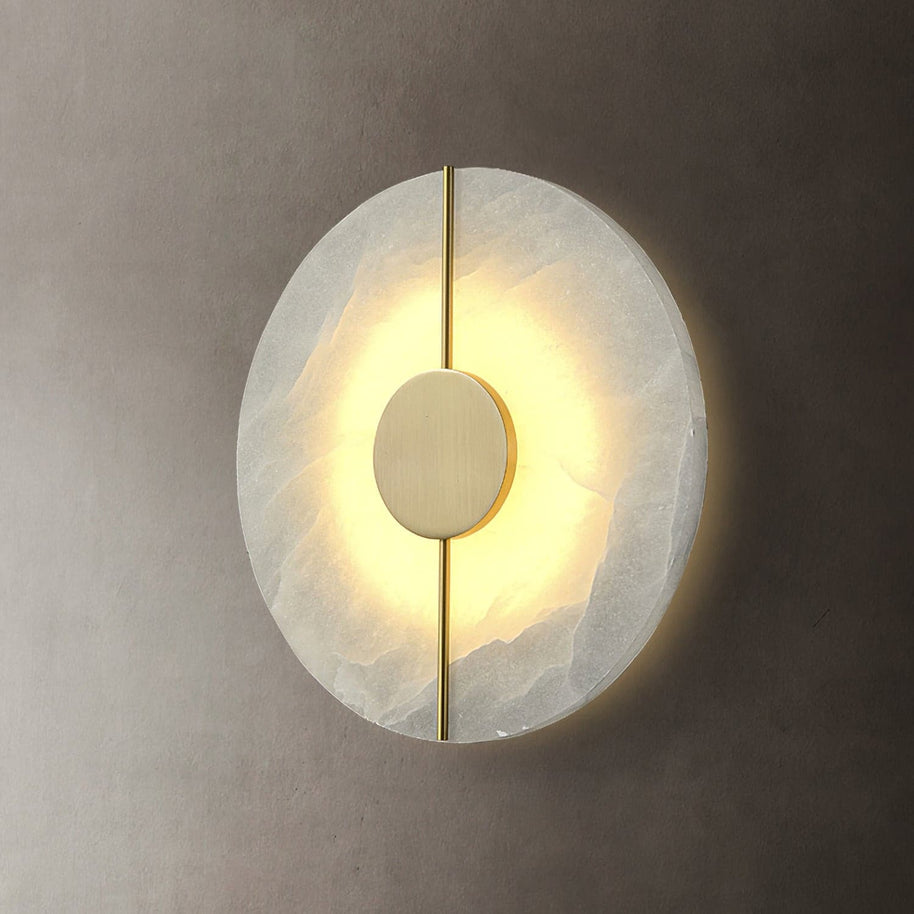 Wall light with a round veined alabaster disc, a slim brass rod and a brass centre disc lit on a plaster wall.