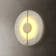 Wall light with a round veined alabaster disc, a slim brass rod and a brass centre disc lit on a plaster wall.