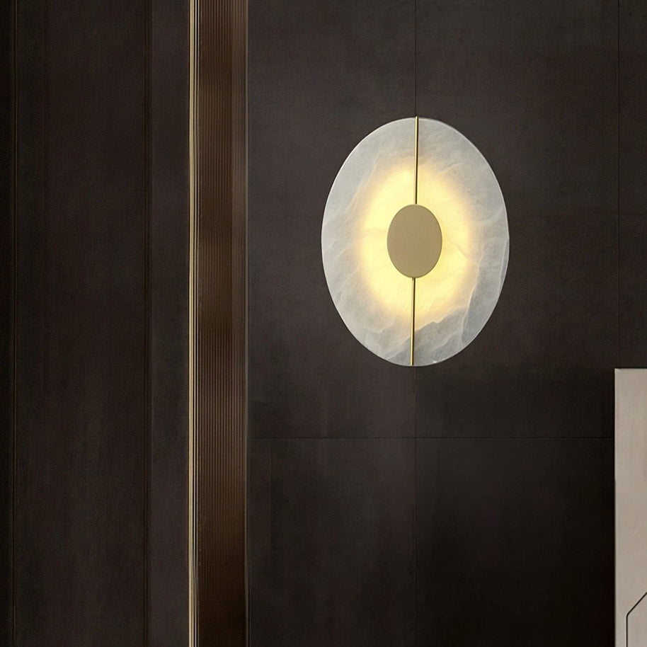 Wall light with a round veined alabaster disc, a slim brass rod and a brass centre disc lit on a wall beside dark panelling.