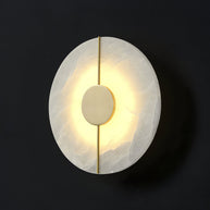 Wall light with a round veined alabaster disc, a slim brass rod and a brass centre disc lit on a dark background, seen close.