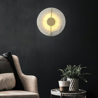 Wall light with a round veined alabaster disc, a slim brass rod and a brass centre disc lit on a dark wall beside an armchair.