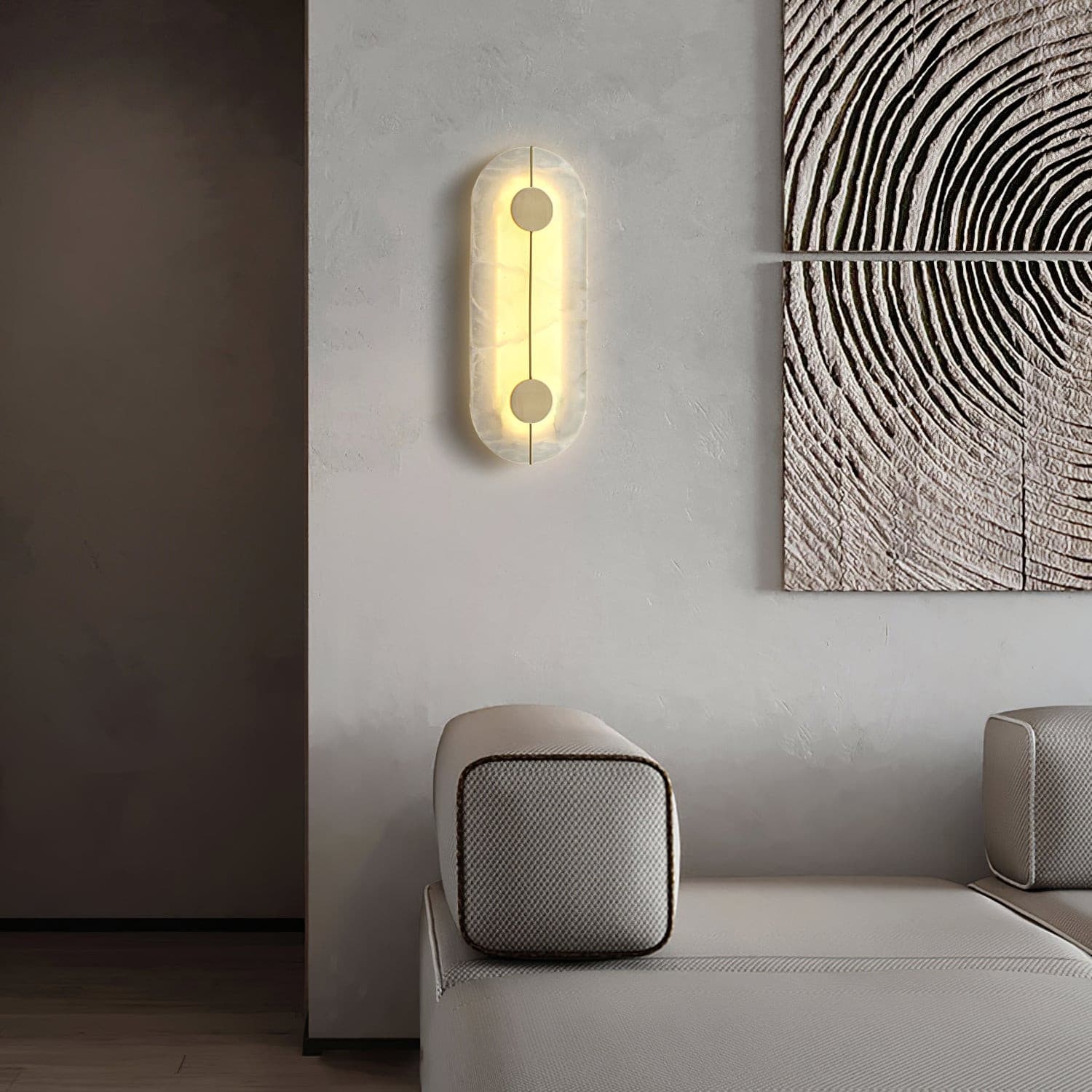 Wall light with a tall rounded alabaster panel, a slim brass rod and two brass discs lit beside a sofa and framed artwork.