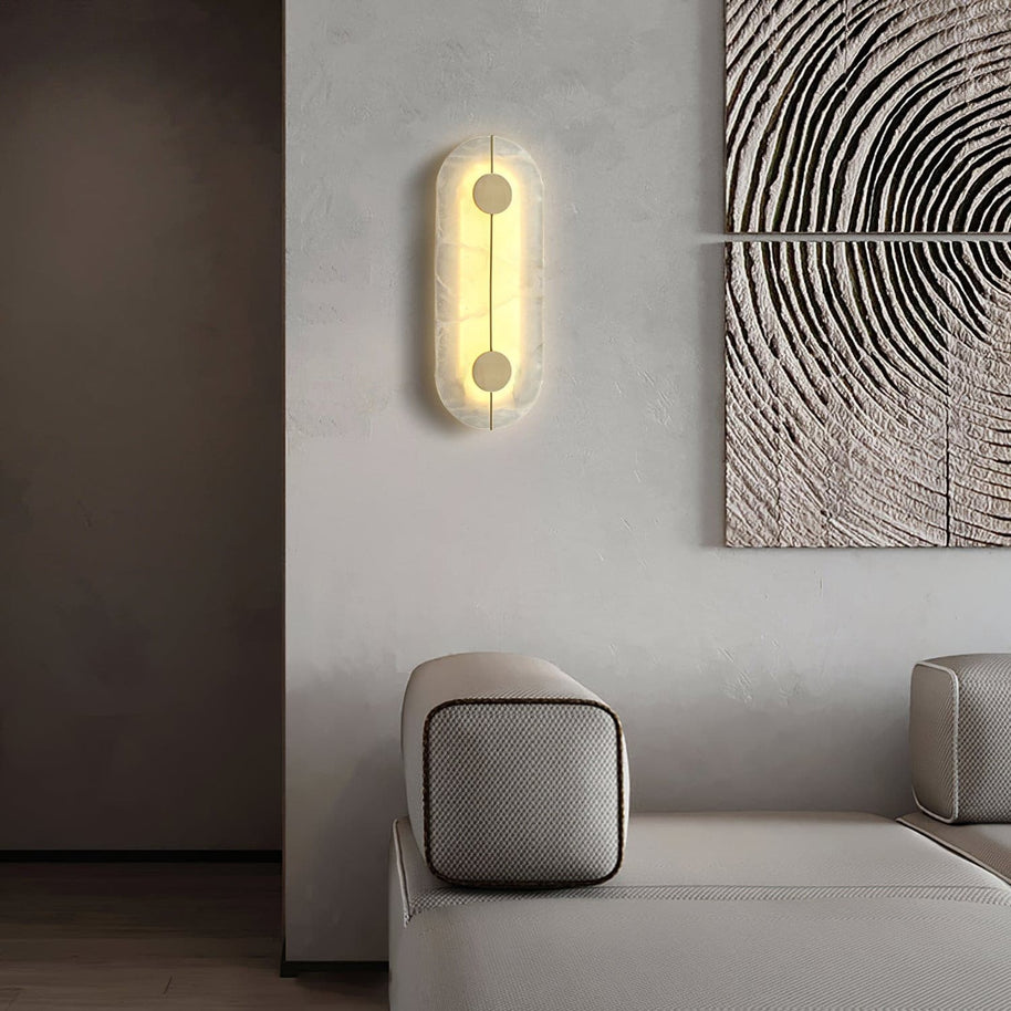 Wall light with a tall rounded alabaster panel, a slim brass rod and two brass discs lit beside a sofa and framed artwork.