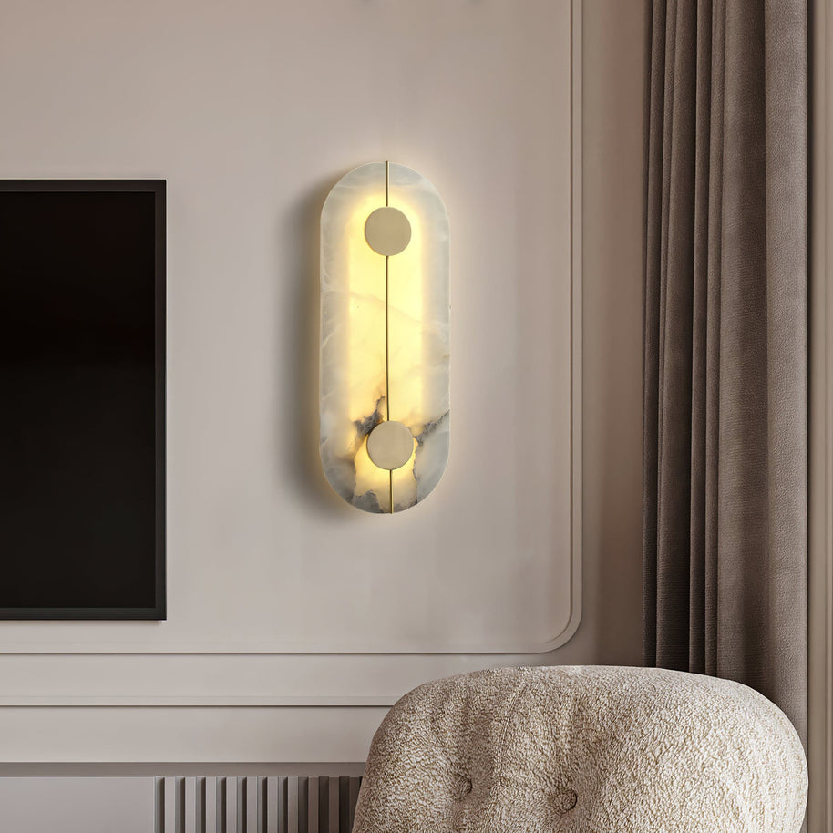 Wall light with a tall rounded alabaster panel, a slim brass rod and two brass discs lit on a panelled wall above an armchair.