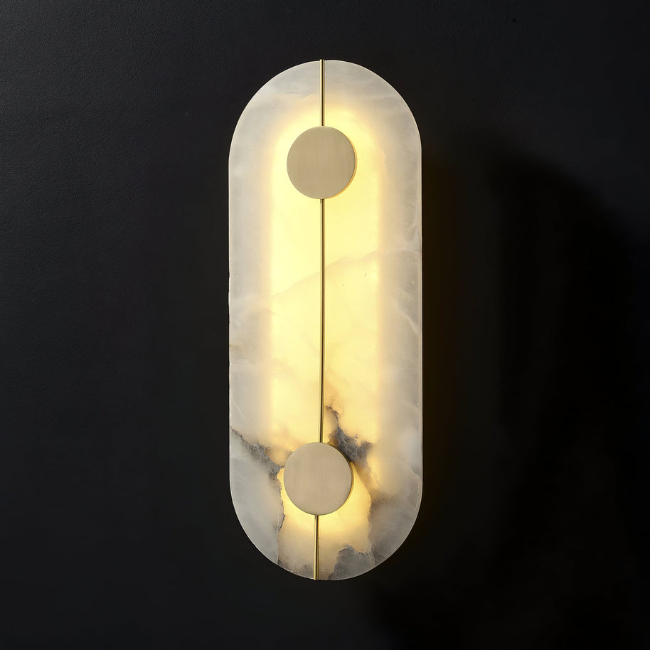Wall light with a tall rounded alabaster panel, a slim brass rod and two brass discs lit on a dark background.