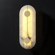Wall light with a tall rounded alabaster panel, a slim brass rod and two brass discs lit on a dark background.