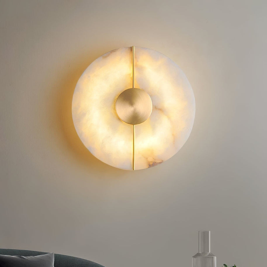 Wall light with a round veined alabaster disc, a slim brass rod and a brass centre disc lit, shown large on a pale wall.