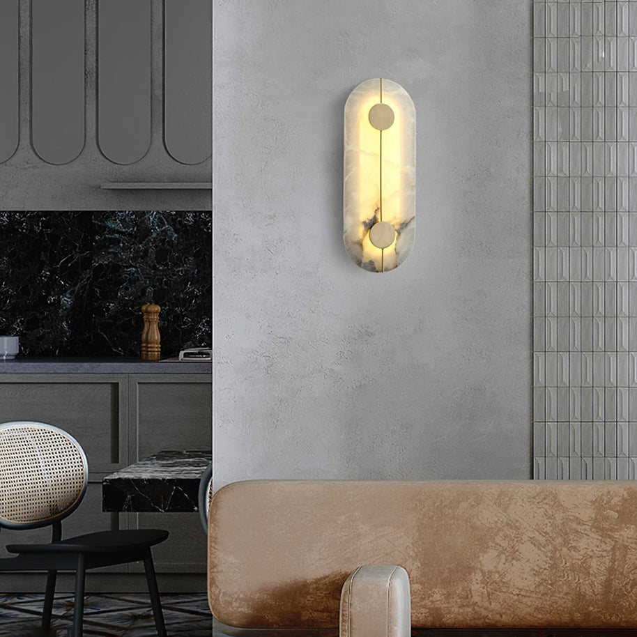 Wall light with a tall rounded alabaster panel, a slim brass rod and two brass discs lit on a plaster wall beside a kitchen.