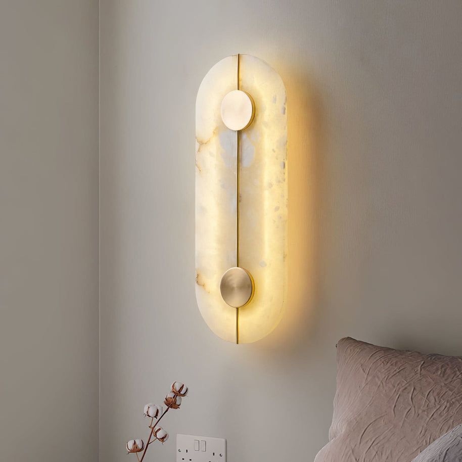 Wall light with a tall rounded alabaster panel, a slim brass rod and two brass discs lit beside a bed.
