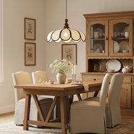 Pendant light with a scalloped beige shade, dark ribs and a turned walnut-coloured wooden top lit above a wooden dining table beside a dresser.