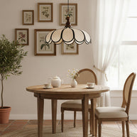 Pendant light with a scalloped beige shade, dark ribs and a turned walnut-coloured wooden top above a round table below framed botanical prints.