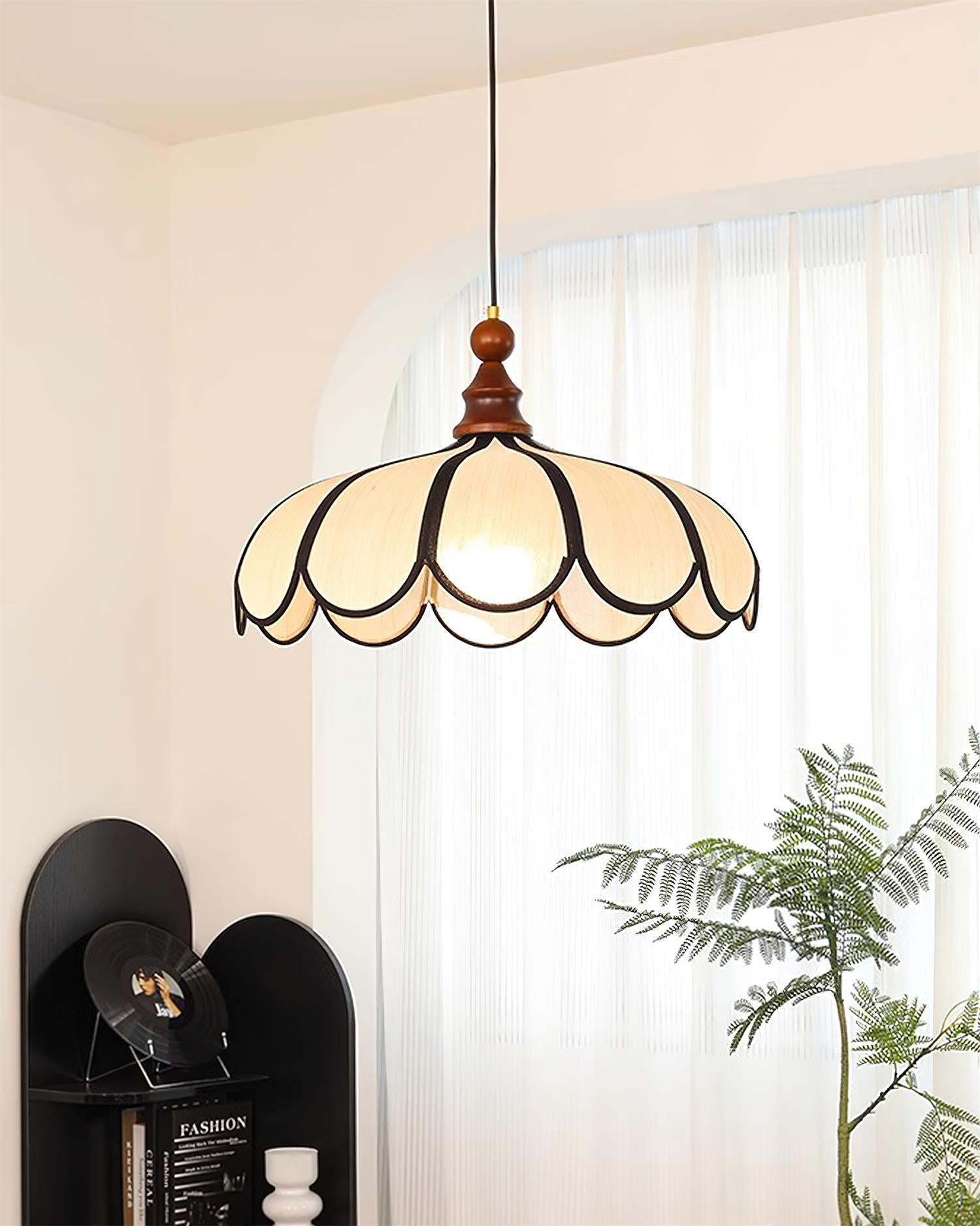 Pendant light with a scalloped beige shade, dark ribs and a turned walnut-coloured wooden top lit beside a curtain and a plant.