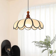 Pendant light with a scalloped beige shade, dark ribs and a turned walnut-coloured wooden top lit beside a curtain and a plant.