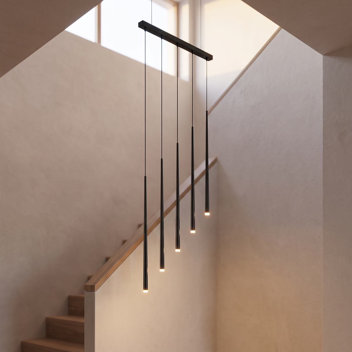 Tirelle chandelier with five slender black rod pendants at staggered drops from one long linear black canopy, lit warm over a plaster stairwell.