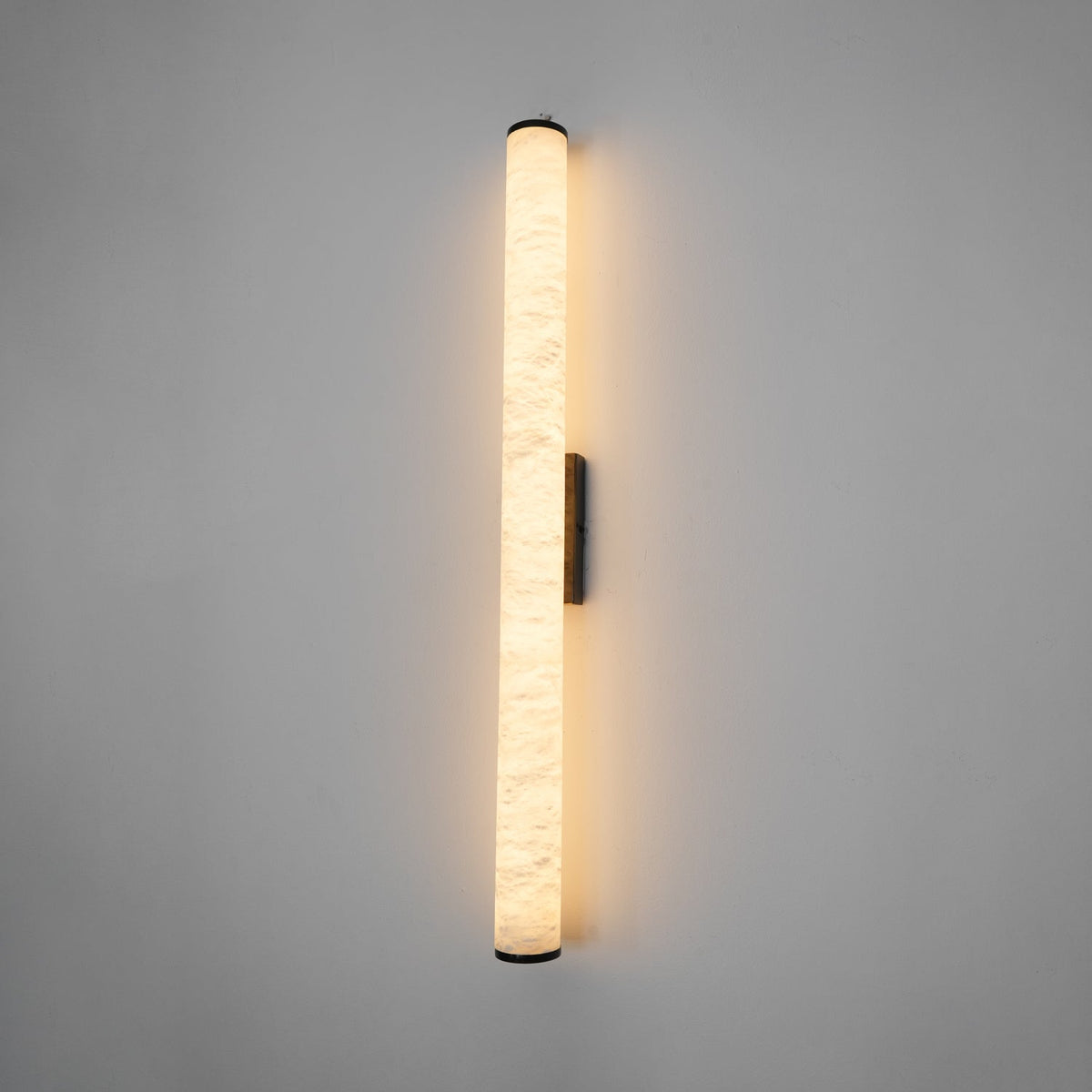 Wall lamp with a long pale veined stone-look tube and a dark bracket, lit, shown long on a grey wall.