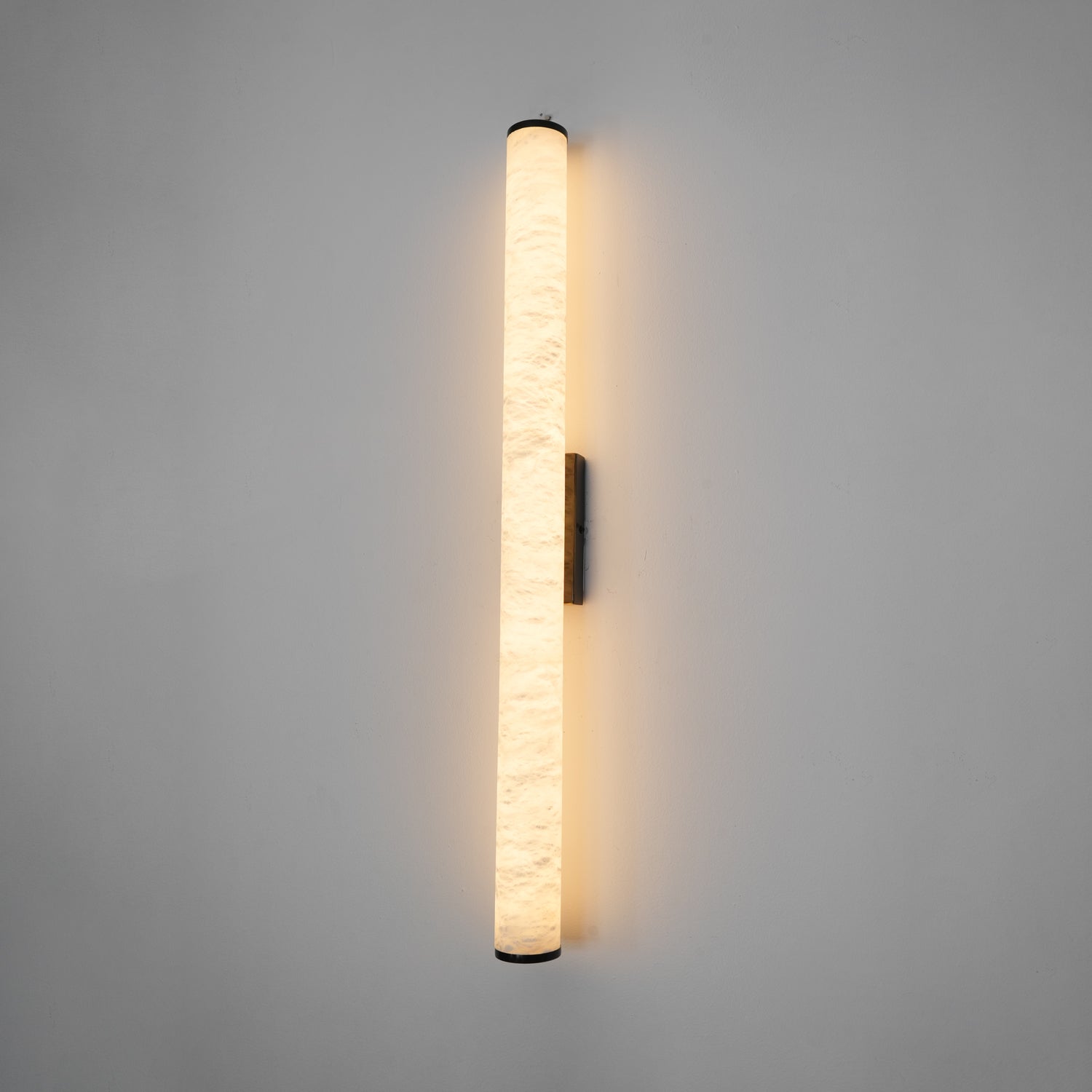 Wall lamp with a long pale veined stone-look tube and a dark bracket, lit, shown long on a grey wall.
