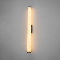 Wall lamp with a long pale veined stone-look tube and a dark bracket, lit, shown long on a grey wall.