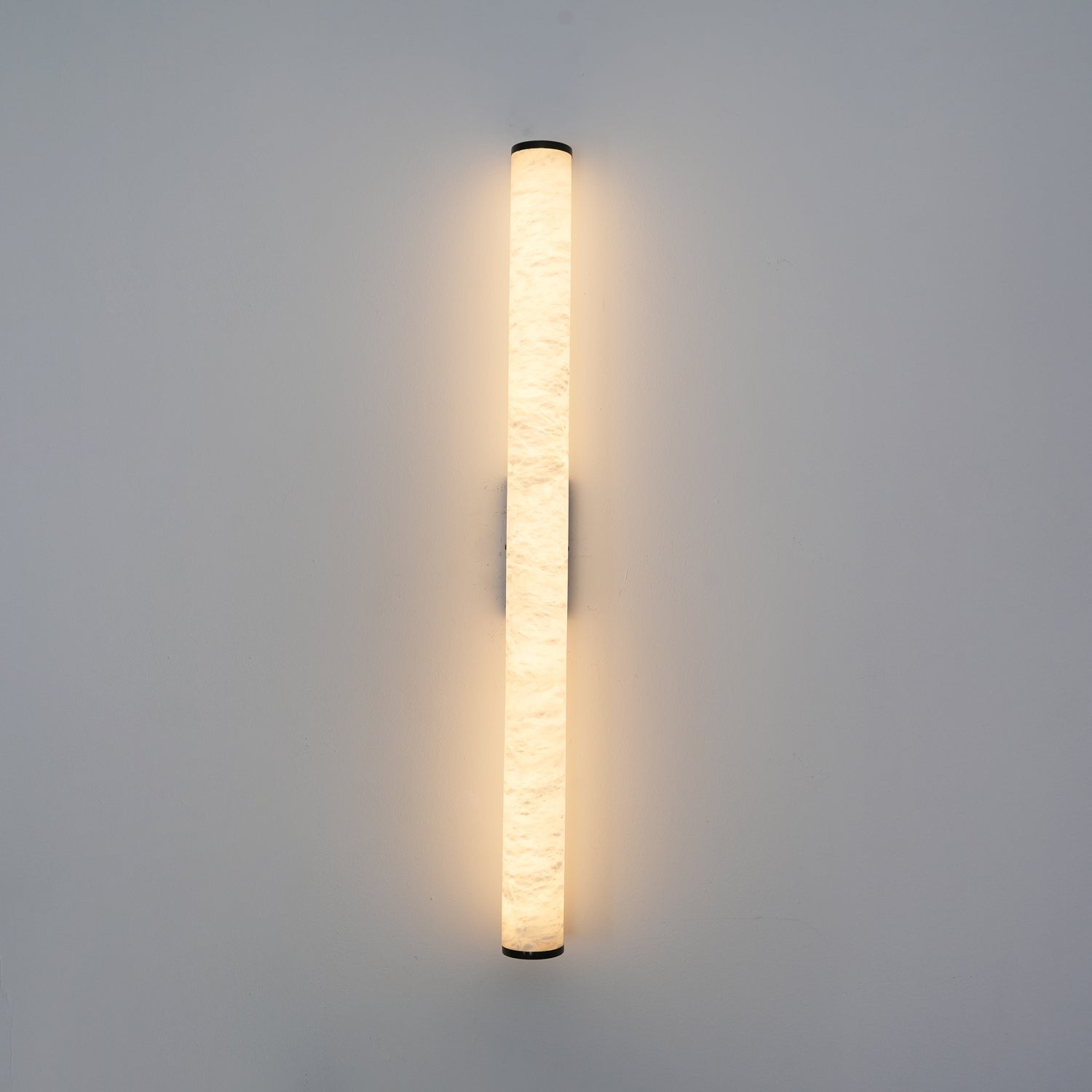 Wall lamp with a long pale veined alabaster tube and a black bracket lit, shown large on a plain wall.