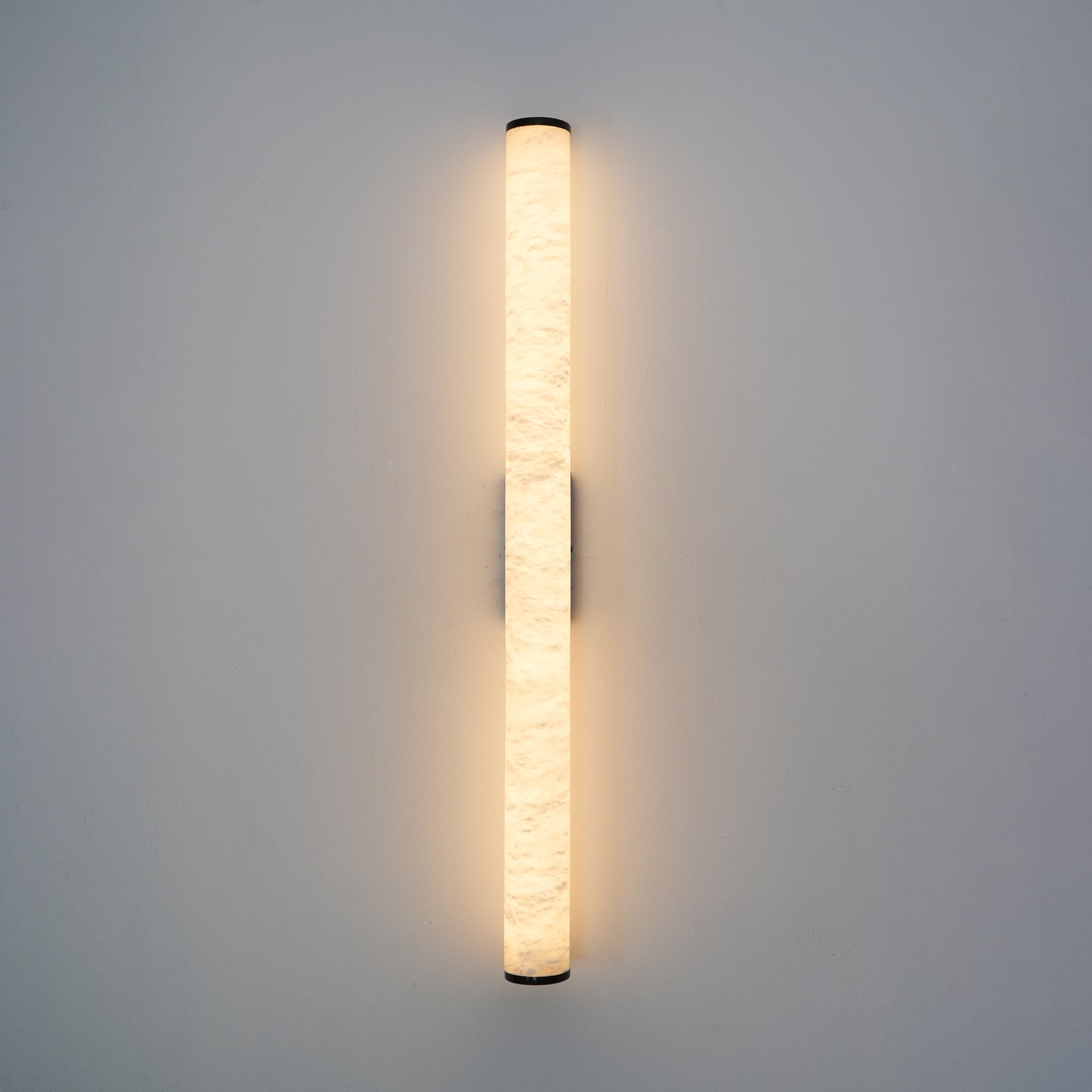 Wall lamp with a long pale veined alabaster tube and a black bracket lit, shown large on a plain wall.