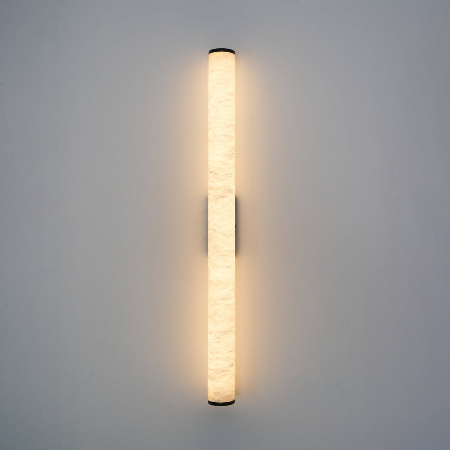 Wall lamp with a long pale veined alabaster tube and a black bracket lit, shown large on a plain wall.