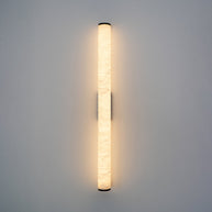 Wall lamp with a long pale veined alabaster tube and a black bracket lit, shown large on a plain wall.