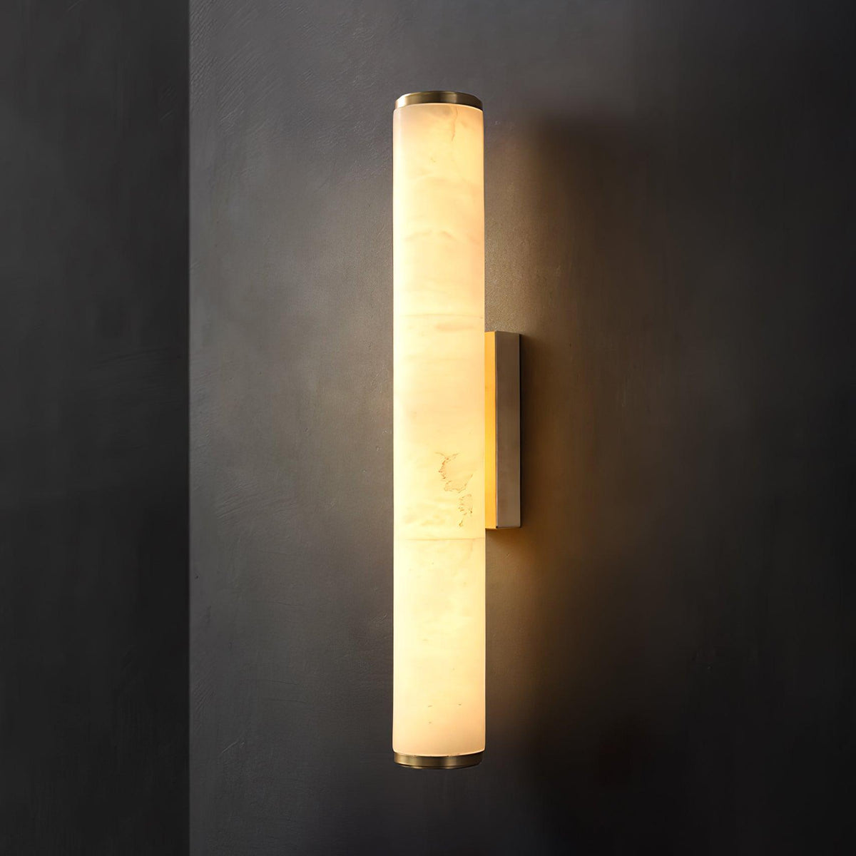 Wall lamp with a long pale veined stone-look tube and a gold-toned bracket, lit, on a dark grey wall.