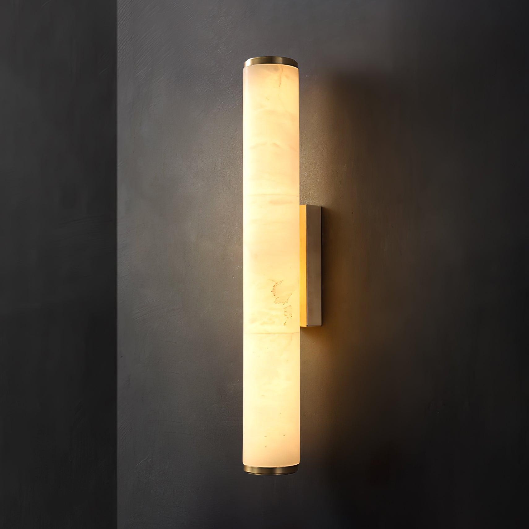 Wall lamp with a long pale veined stone-look tube and a gold-toned bracket, lit, on a dark grey wall.